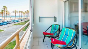 A balcony with two colorful chairs, a glass door, and a view of a parking area with palm trees and a beach. - Roses Holiday Center by Interhome (Roses)