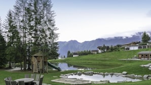 Et udendørs område ved søen med et trælegeland, picnicborde og en dam.