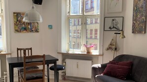 Een gezellige woonkamer met een houten eettafel, een bank, en een ronde salontafel. Er is een raam met uitzicht op gebouwen, een radiator, en diverse wanddecoraties.