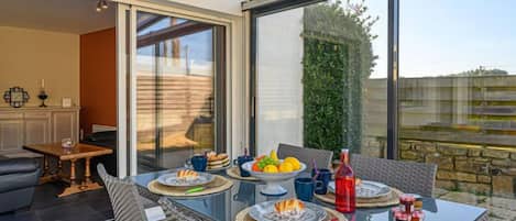 A modern dining area with a glass table, wicker chairs, and a view of the garden through large windows.