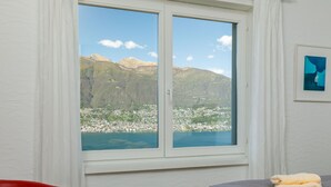Una habitación con una cama, una silla roja, un ventanal con vista a la montaña y un bolso pequeño en el suelo.