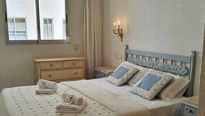 A neatly made bed with pillows, a wooden dresser, a nightstand, and a wall-mounted light fixture. - Marysol by Interhome (Altea)
