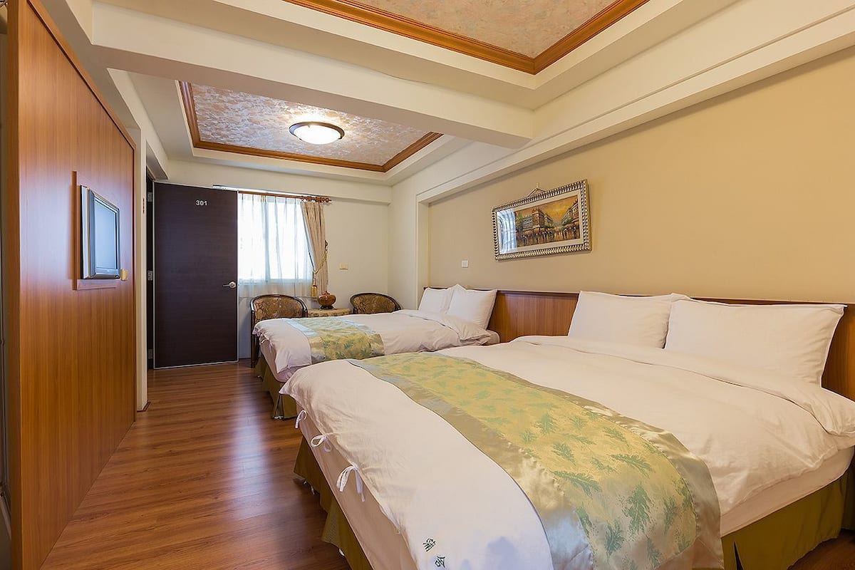 A hotel room with two beds, a wooden floor, a ceiling with a recessed light, a door, and a framed picture on the wall.