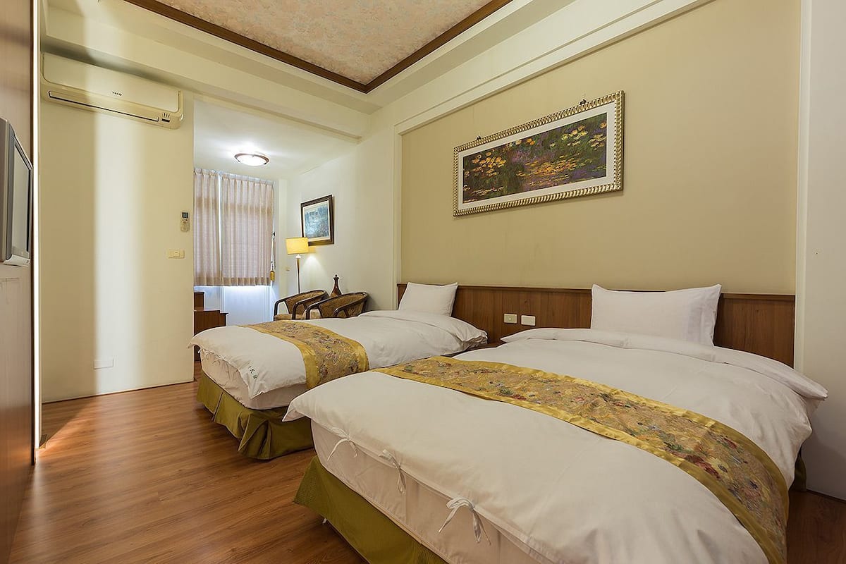 A hotel room with two beds, a wooden floor, a painting on the wall, and a lamp.