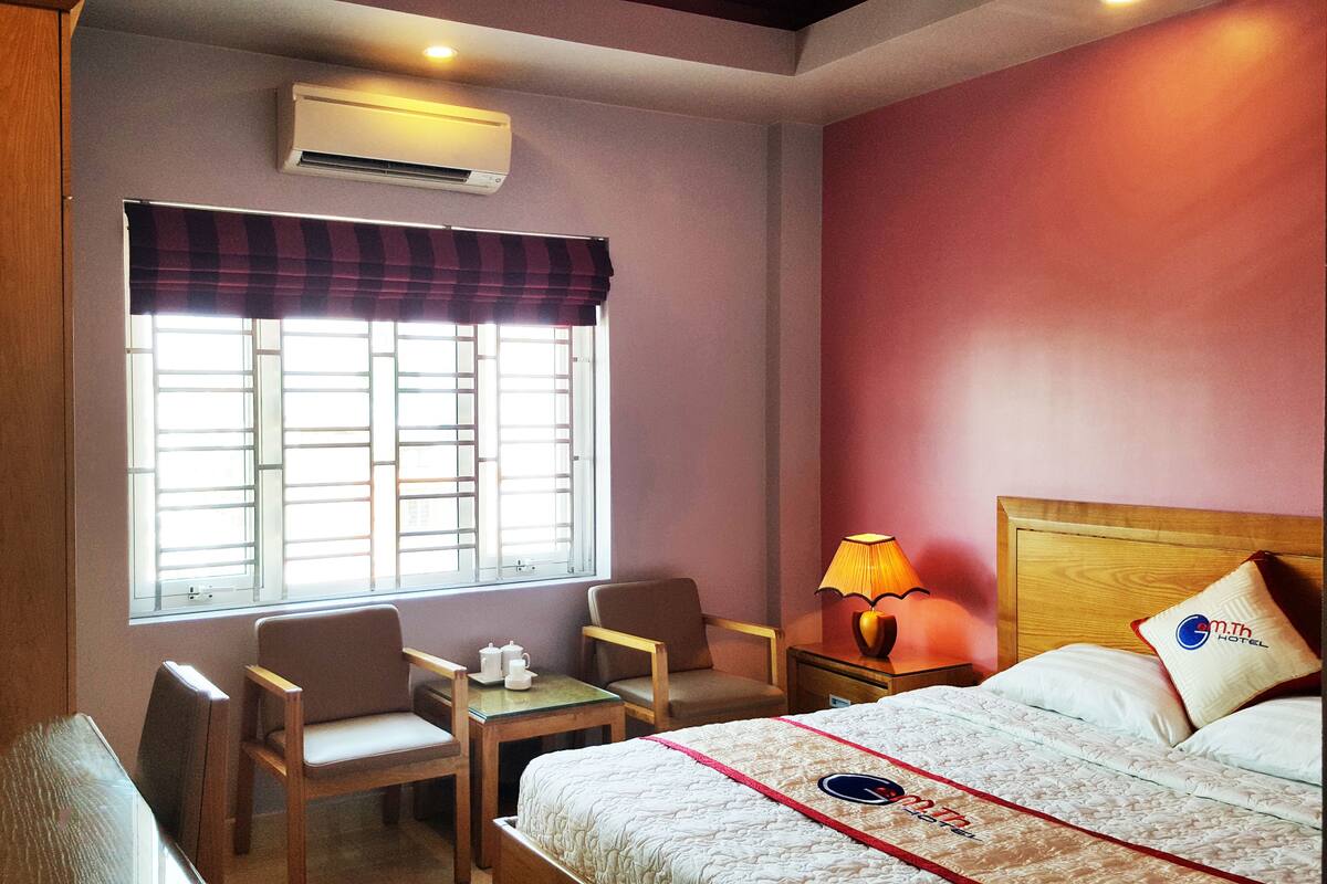 A hotel room with a bed, two chairs, a small table with a teapot, a lamp, and an air conditioner.