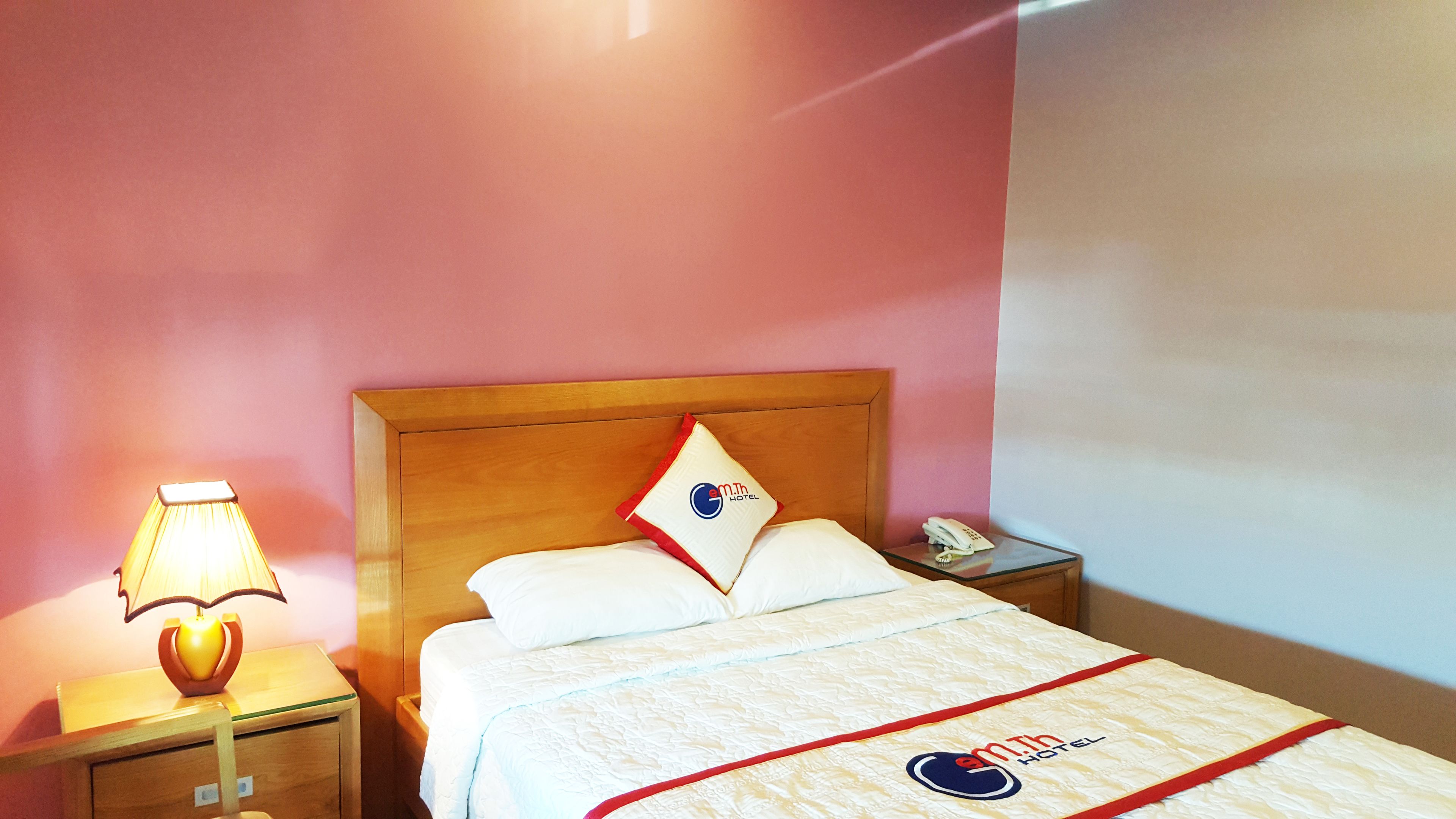 Une chambre d’hôtel avec une tête de lit en bois, un lit avec une literie blanche et un oreiller rouge et bleu, une lampe de chevet et un téléphone sur une table de chevet.