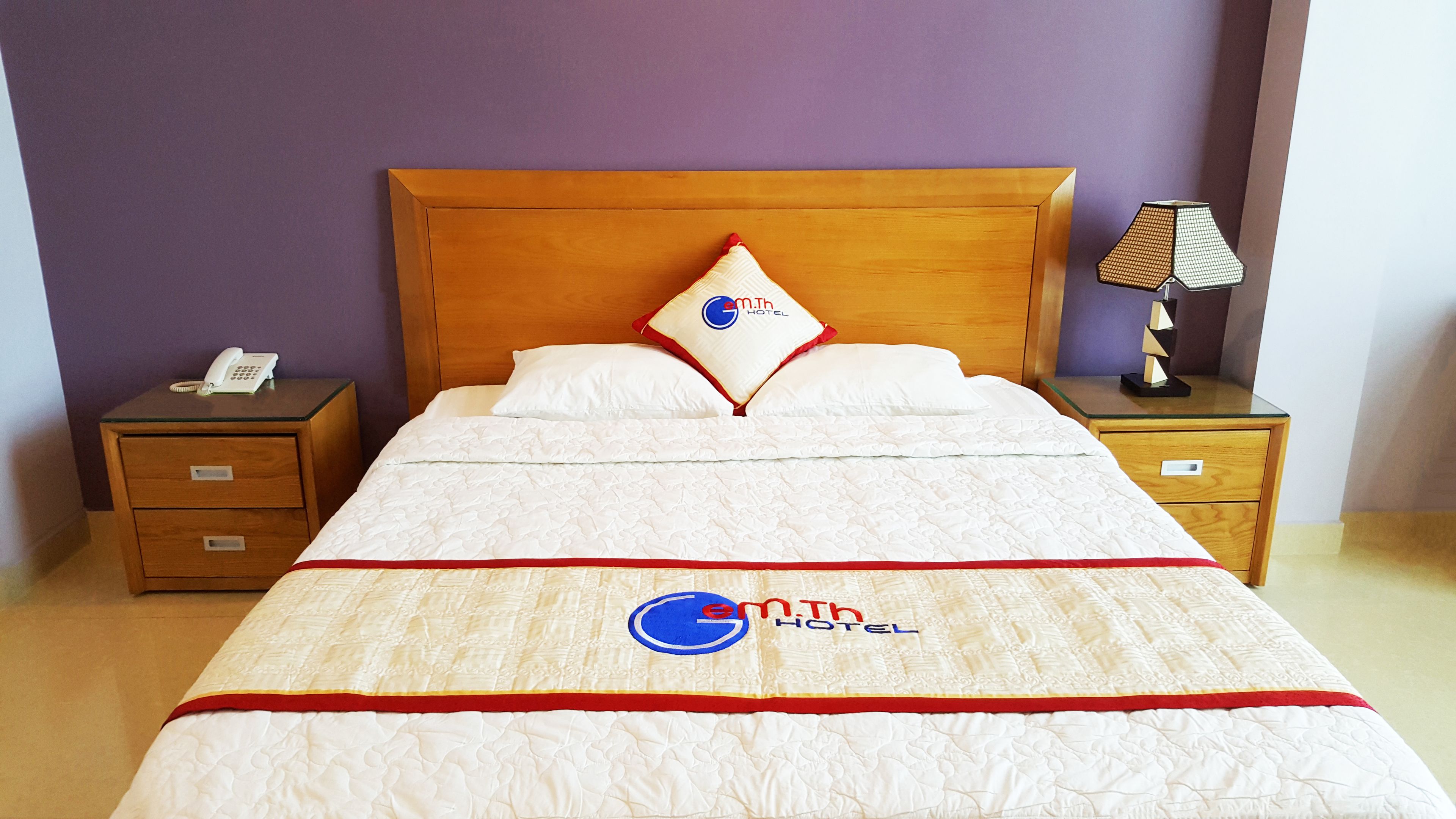 A hotel room with a wooden headboard, a bed with a white quilt and red trim, a pillow with a logo, a bedside table with a lamp, and a phone on the nightstand.