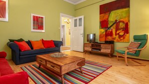 Een woonkamer met een blauwe bank, rode kussens, een houten salontafel, een gestreept vloerkleed, een televisie op een houten kast en een groot abstract schilderij aan de muur.