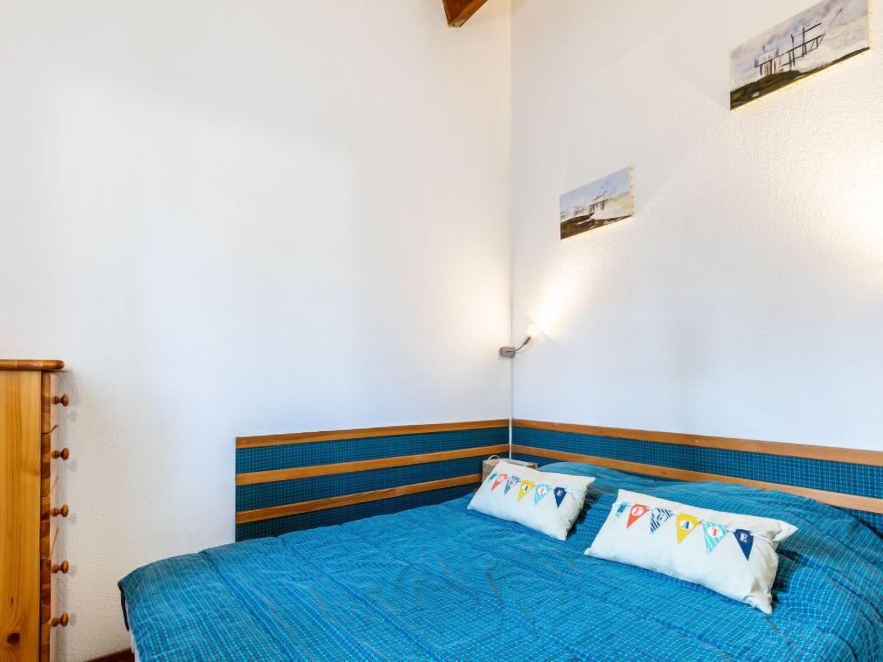 A bedroom with a bed, pillows, a wall-mounted light, and a small painting.