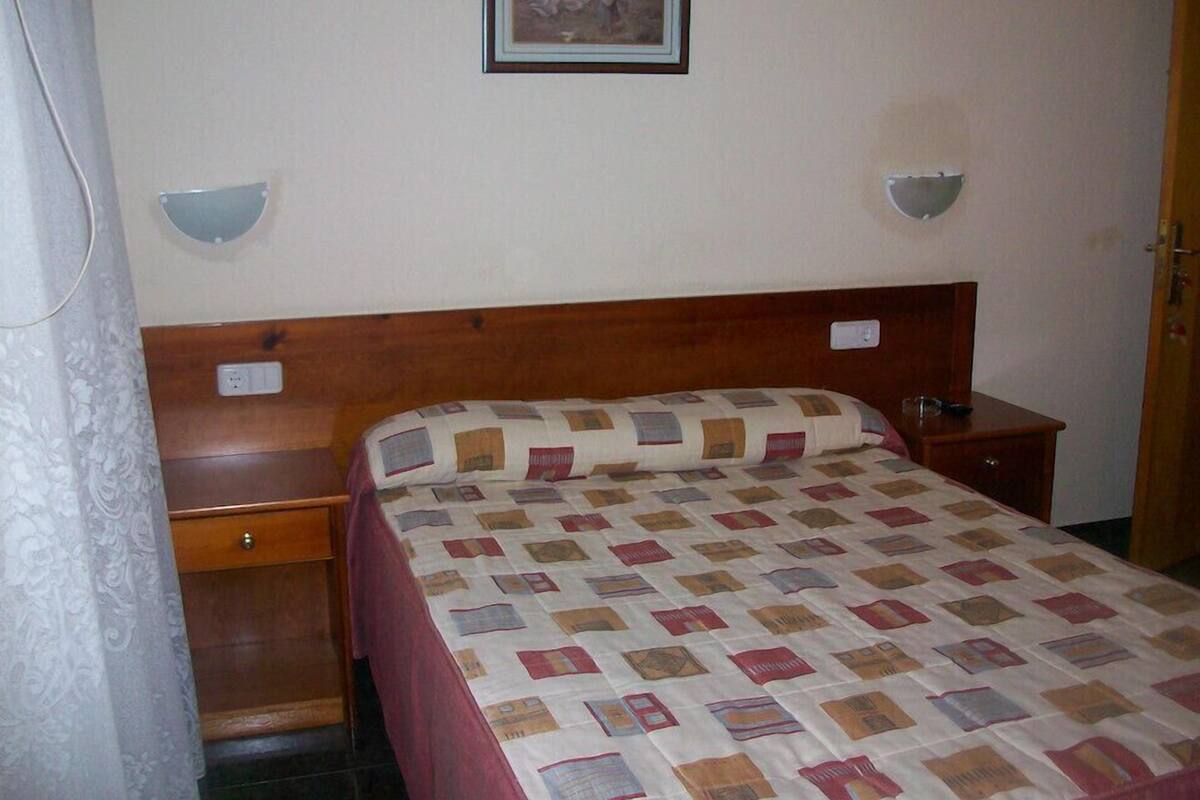 A bedroom with a wooden headboard, a quilted bedspread, a bedside table, a nightstand, and a wall-mounted lamp.