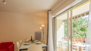 Uma área de refeições com uma mesa posta para dois, uma cadeira vermelha e vista para o pátio externo através de grandes portas de vidro deslizantes.