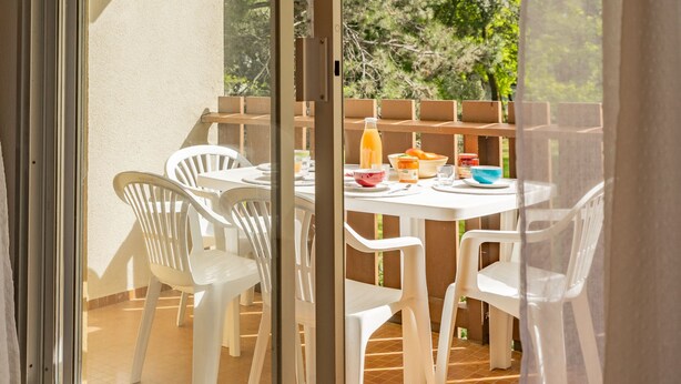 Um pátio com uma mesa posta para o café da manhã, cadeiras de plástico branco e vista para árvores.