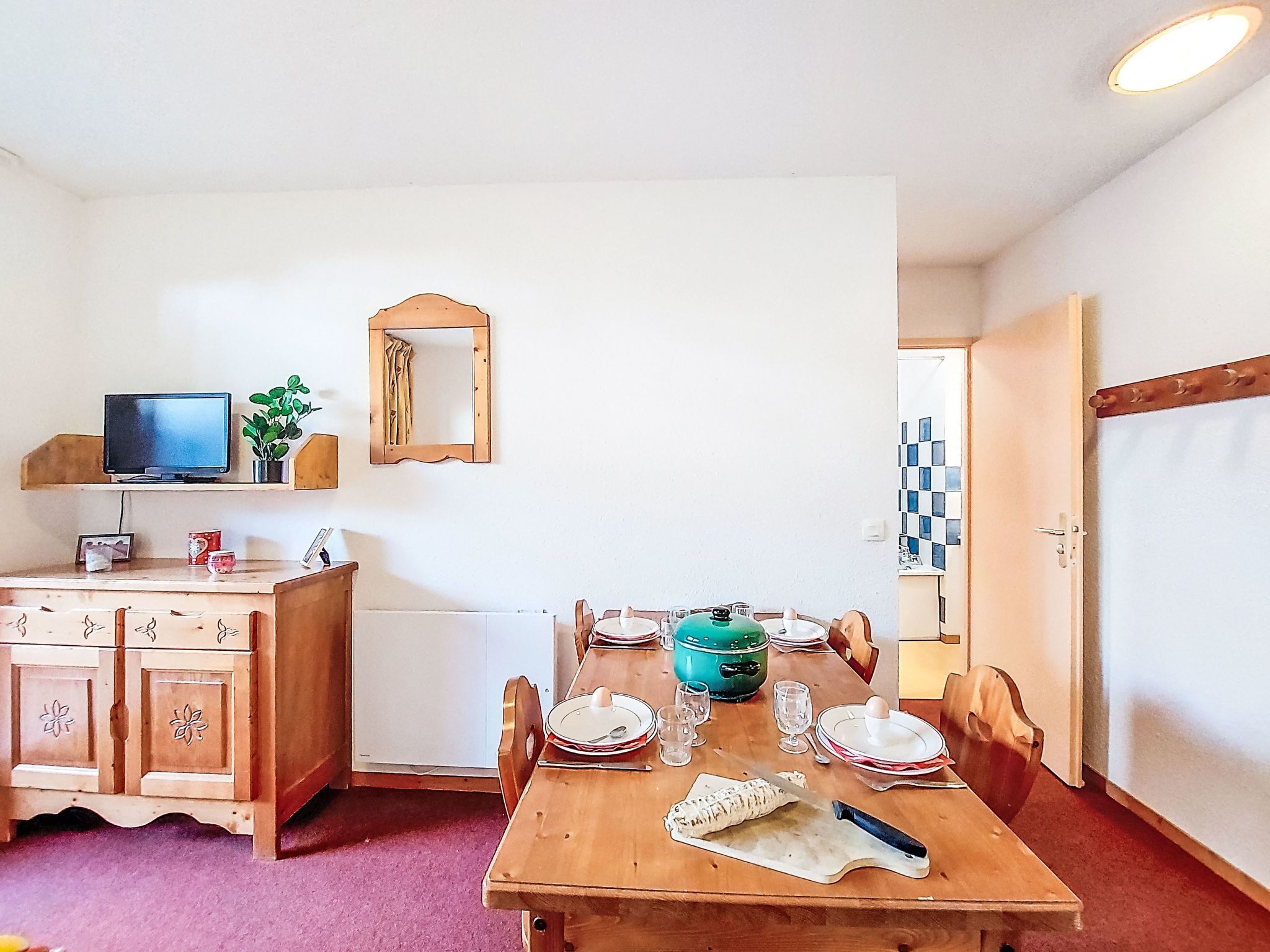 Un comedor con una mesa de madera preparada para cuatro personas, un televisor en un estante, un espejo y un radiador.