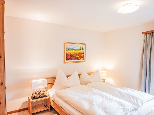 A hotel room with a bed, two bedside tables, a nightstand, a lamp, a painting on the wall, and a window with curtains.
