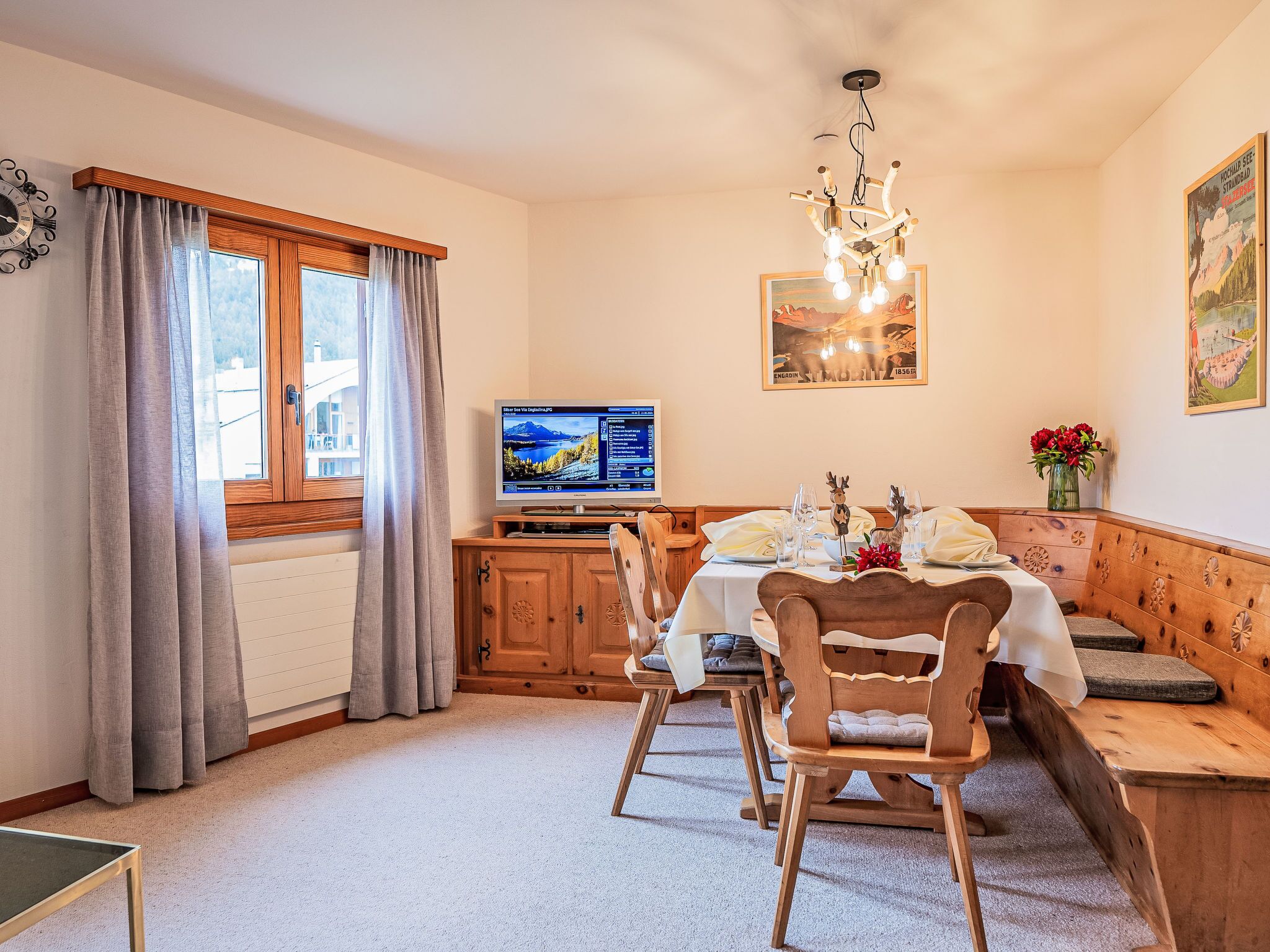 Un coin repas avec une table en bois dressée pour deux personnes, un téléviseur posé sur un meuble en bois et une fenêtre avec des rideaux.