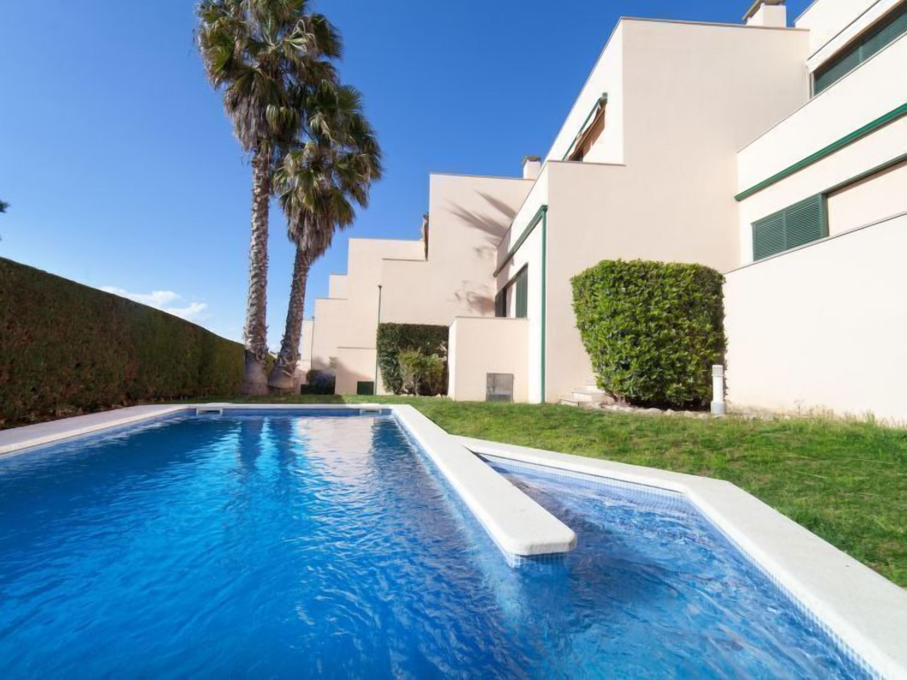 Un área de piscina moderna con una piscina azul cristalina, palmeras y un edificio blanco con detalles verdes.