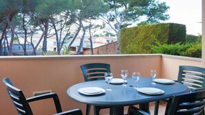 Et rundt bord med seks stole, hver med en tallerken og et glas, placeret på en udendørs terrasse med udsigt til træer og bygninger.