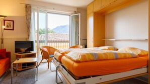 A hotel room with a bed, a TV, a chair, and a view of the mountains. - Residenza Corallo by Interhome (Ascona)