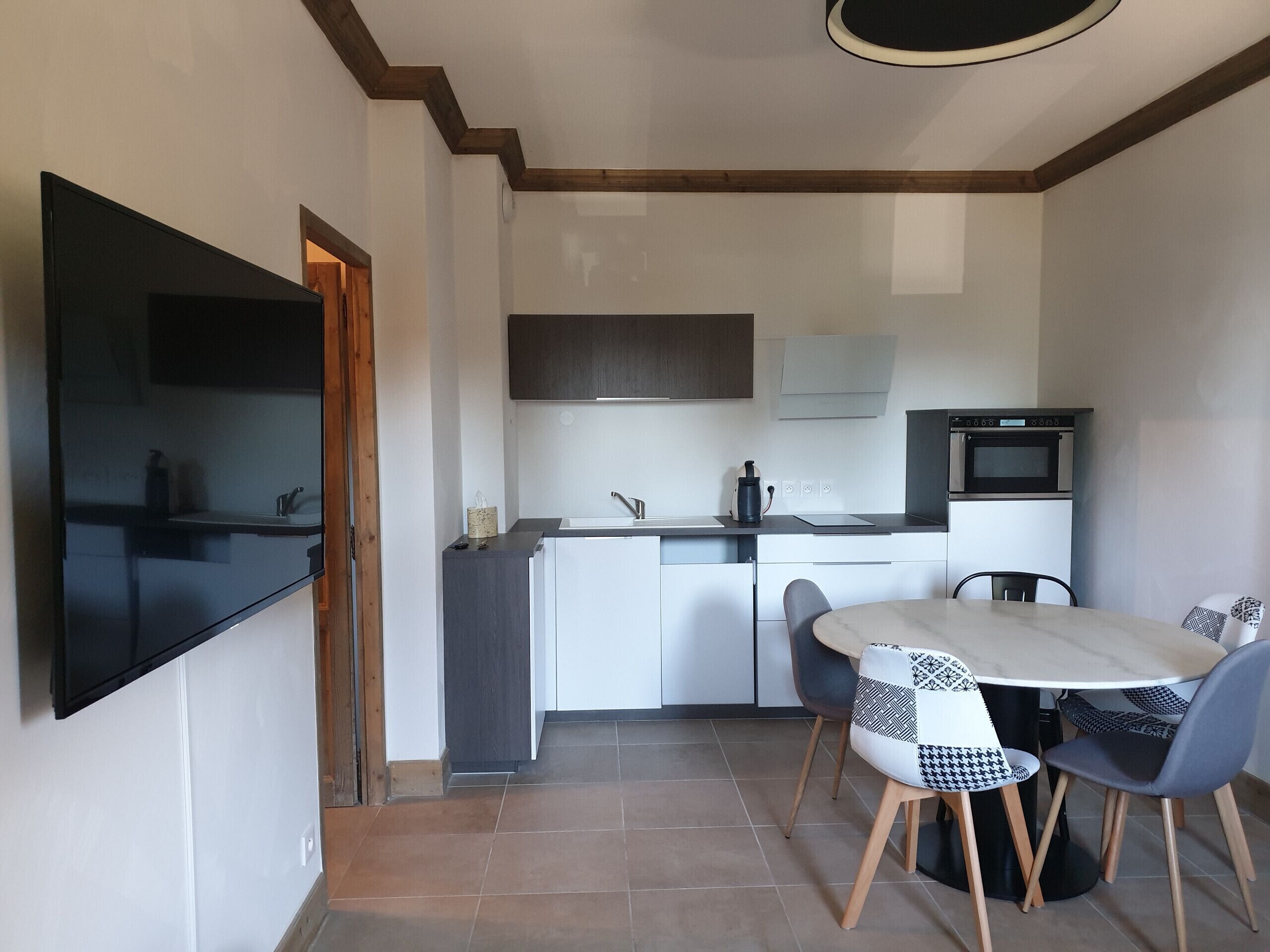 A compact kitchen with a dining area, featuring a flat-screen TV mounted on the wall, a microwave, and a sink.