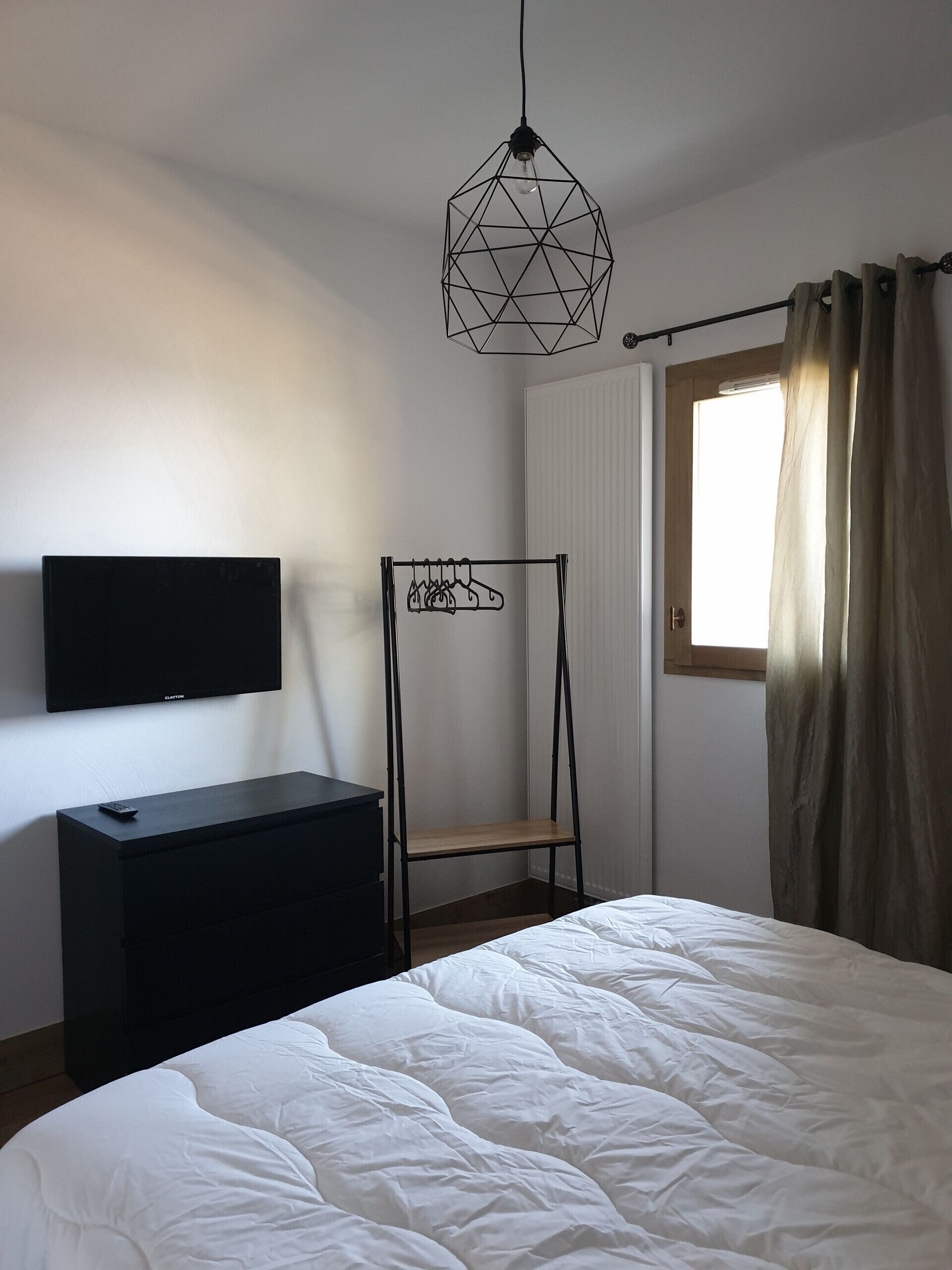 A bedroom with a bed, a TV on a dresser, a hanging light fixture, and a window with curtains.