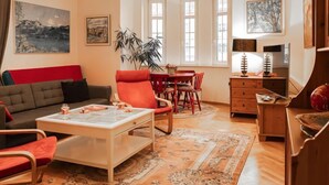 Ein Wohnzimmer mit Sofa, Sesseln, einem Couchtisch, einem Esstisch mit Stühlen, einer Lampe und Bildern an den Wänden.