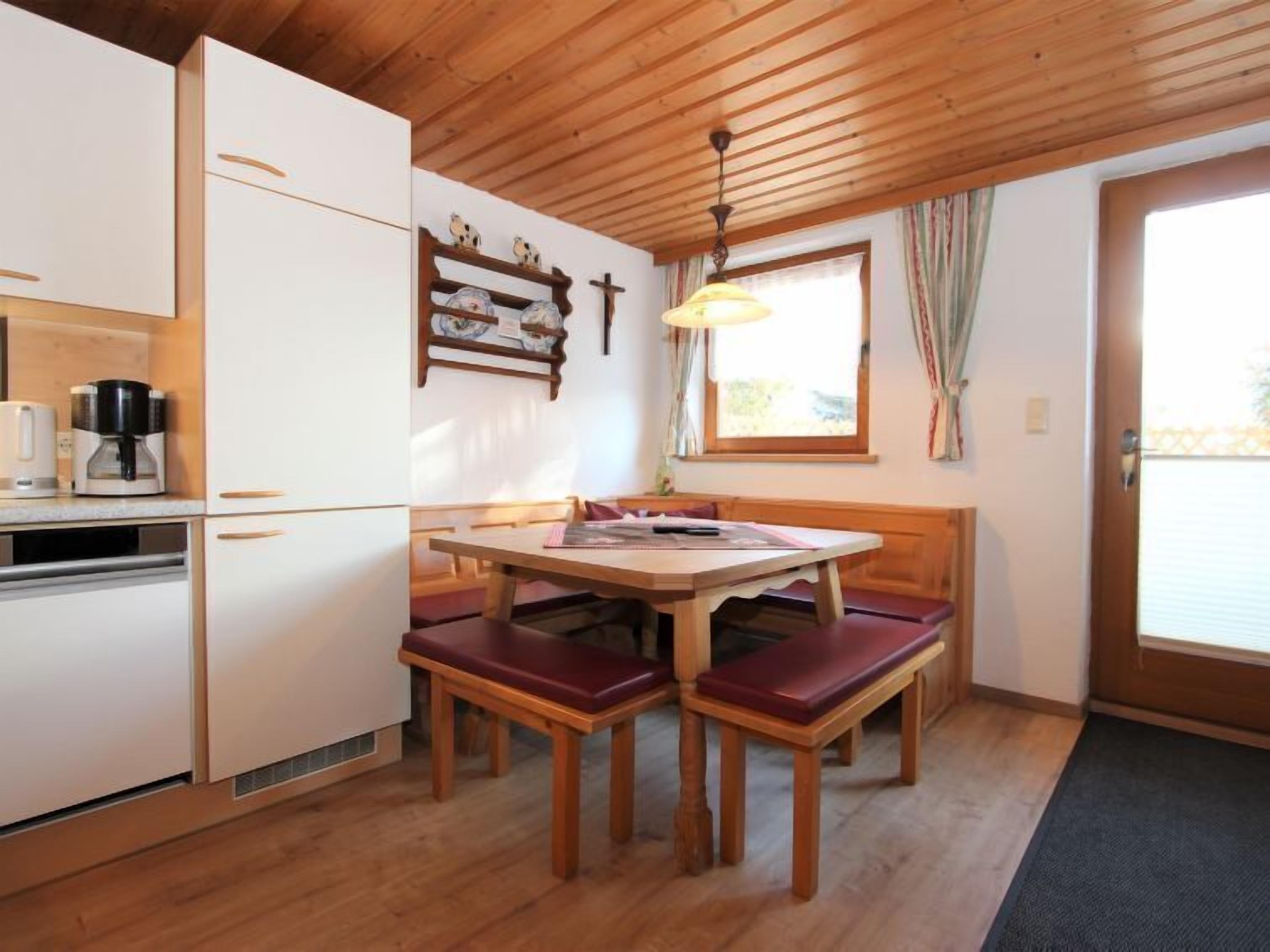 A compact kitchen with white cabinets, a wooden dining table with benches, a coffee maker, and a hanging light fixture.