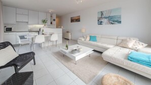 A modern living room with a sofa, coffee table, and chairs. There is a kitchen area with white cabinets and a dining table.