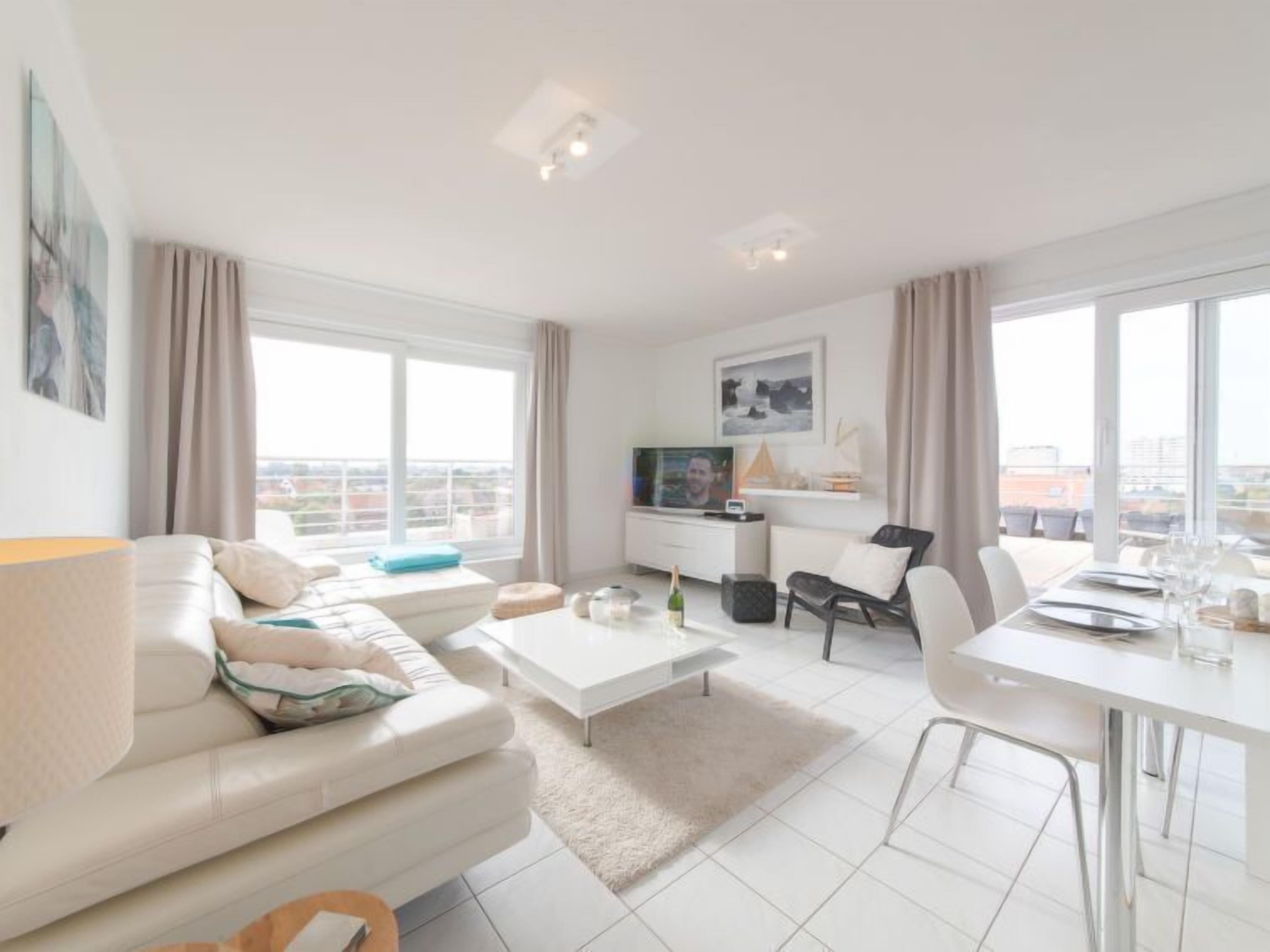 Una sala de estar moderna con sofá, mesa de centro, mesa de comedor y sillas. Hay un televisor sobre un mueble, una ventana con cortinas y vistas a la ciudad.