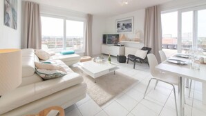 A modern living room with a sofa, coffee table, dining table, and chairs. There is a TV on a stand, a window with curtains, and a city view.