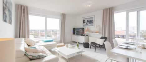Una sala de estar moderna con sofá, mesa de centro, mesa de comedor y sillas. Hay un televisor sobre un mueble, una ventana con cortinas y vistas a la ciudad.