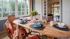Un comedor con una gran mesa redonda dispuesta para cuatro personas, rodeada de sillas de madera con cojines rojos. Un jarrón con flores está sobre la mesa, y la habitación tiene ventanas grandes con vistas a zonas verdes al exterior.