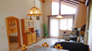 A cozy living room with a sofa, a table with floral tablecloth, wooden cabinets, a television, and a large window with curtains. - Metropol A 13 by Interhome (Grächen)