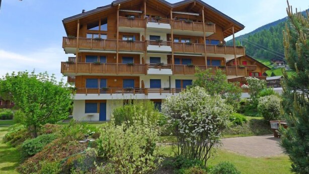 A multi-story wooden hotel with balconies, surrounded by greenery and a mountainous backdrop. - Metropol A 13 by Interhome (Grächen)