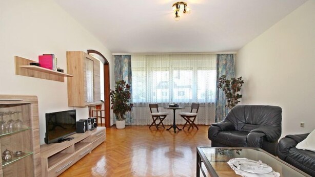 A living room with a wooden floor, a black leather sofa, a glass coffee table, a TV, and a dining area with two chairs and a table.