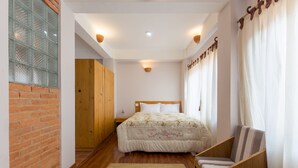 A bedroom with a wooden bed, a nightstand, a chair, a brick wall, and a window with a view. - Team Yala Inn (Lalitpur)