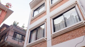 A multi-story building with brick and white exterior walls, multiple windows with white frames, and a visible balcony with a metal railing. - Team Yala Inn (Lalitpur)
