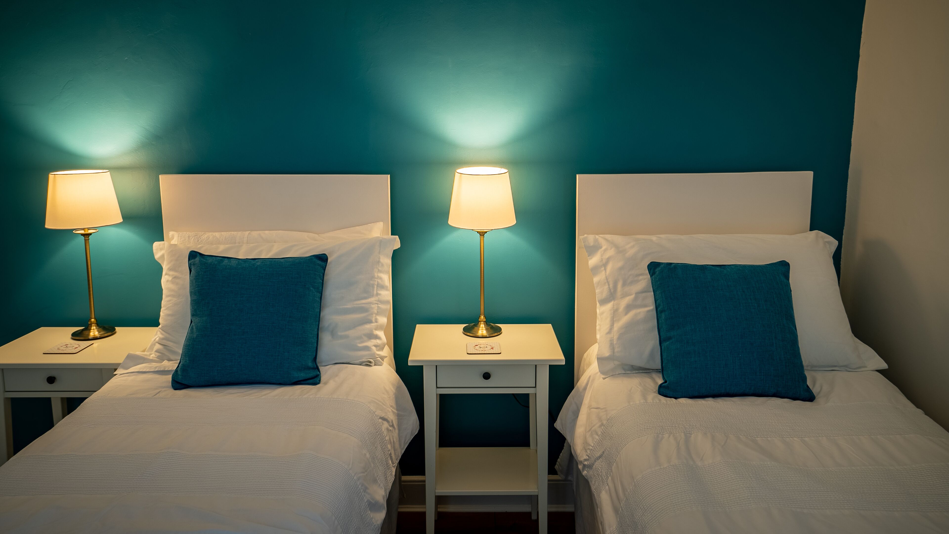 A hotel room with two single beds, each with a white headboard, a teal pillow, and a bedside table with a lamp.