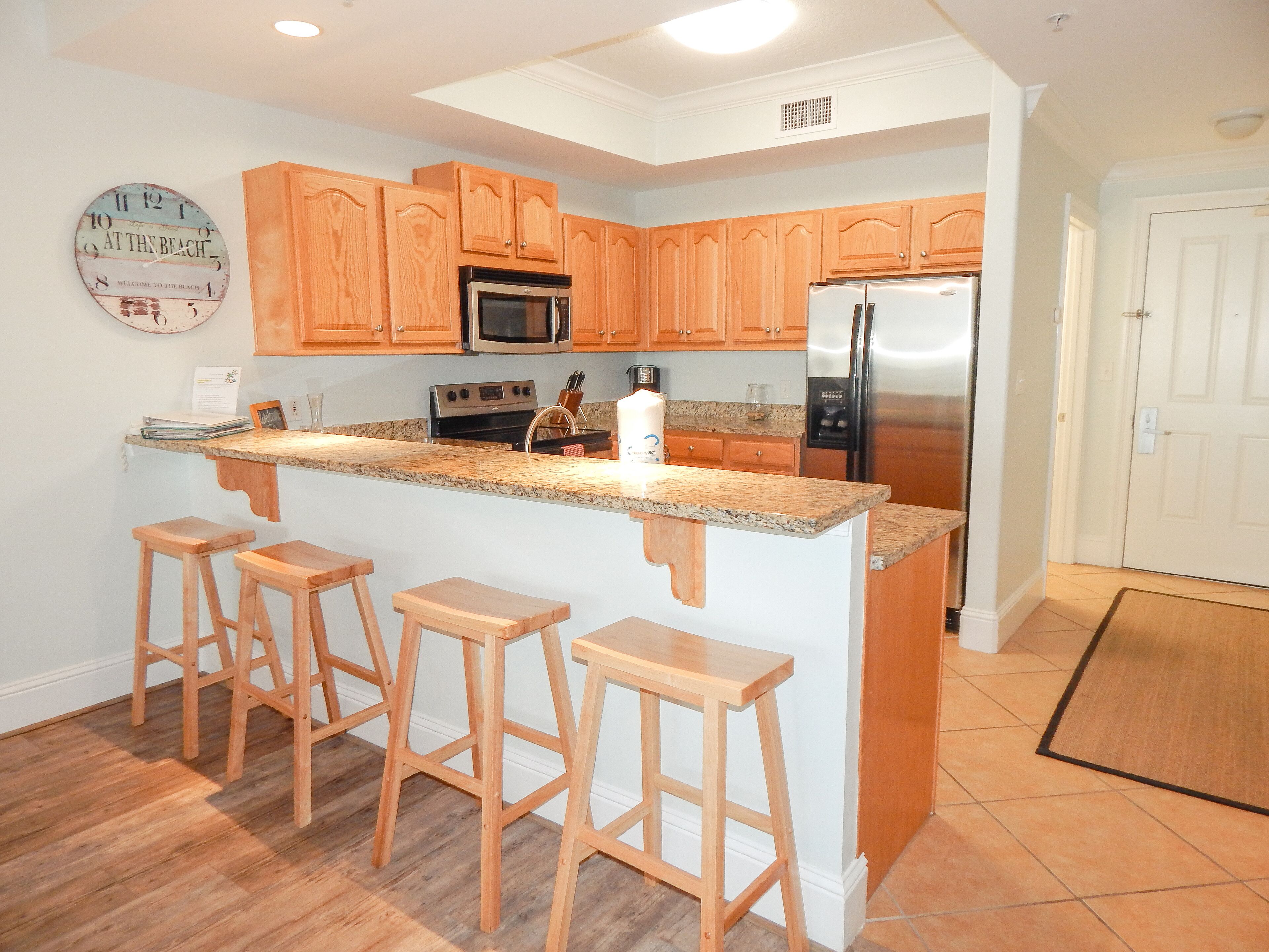 A kitchen with wooden cabinets, a stainless steel refrigerator, a microwave, and a granite countertop. There are three wooden stools and a small island with a sink.