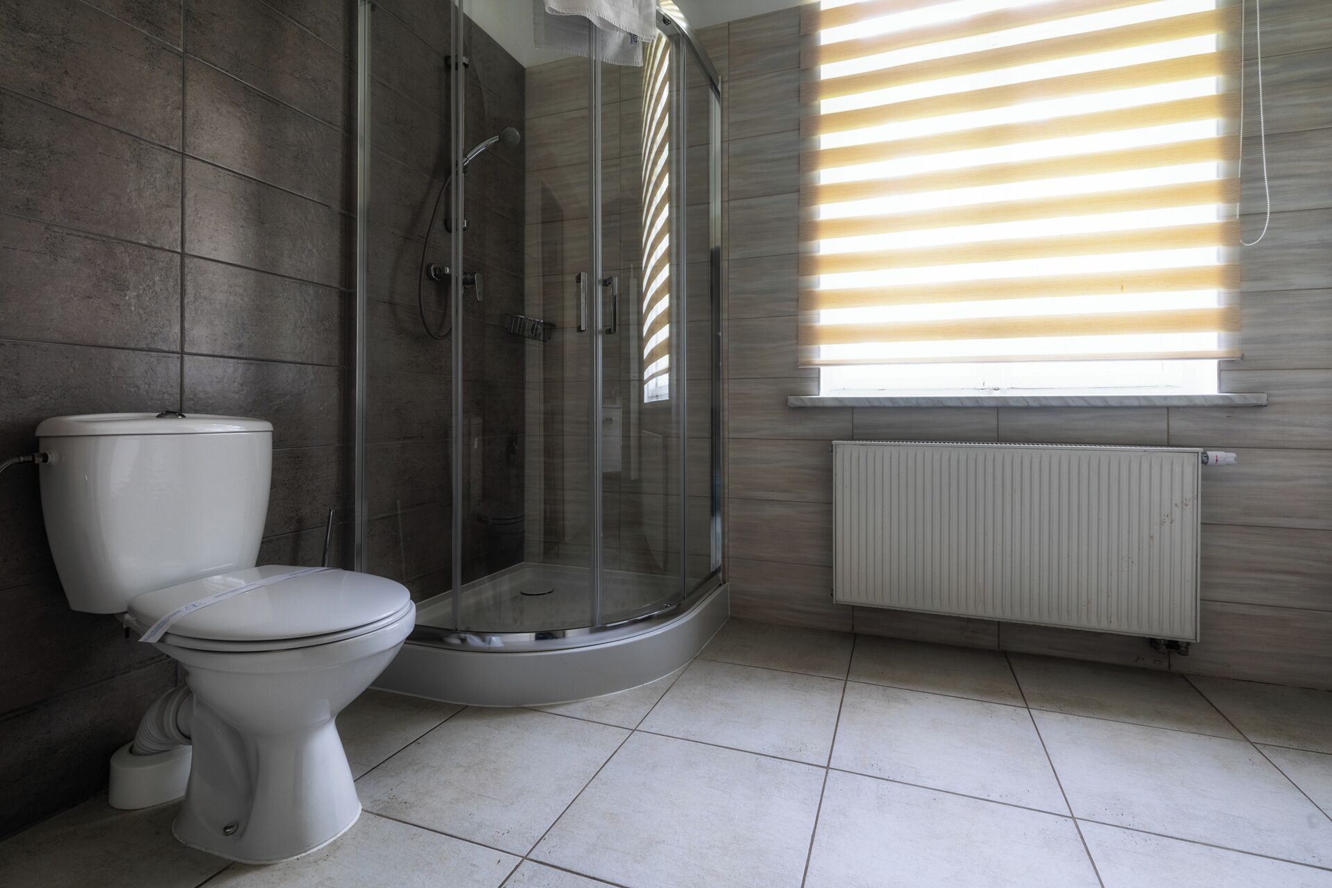 Une salle de bain comprenant des toilettes, une douche fermée par une paroi de verre et une fenêtre avec des stores.