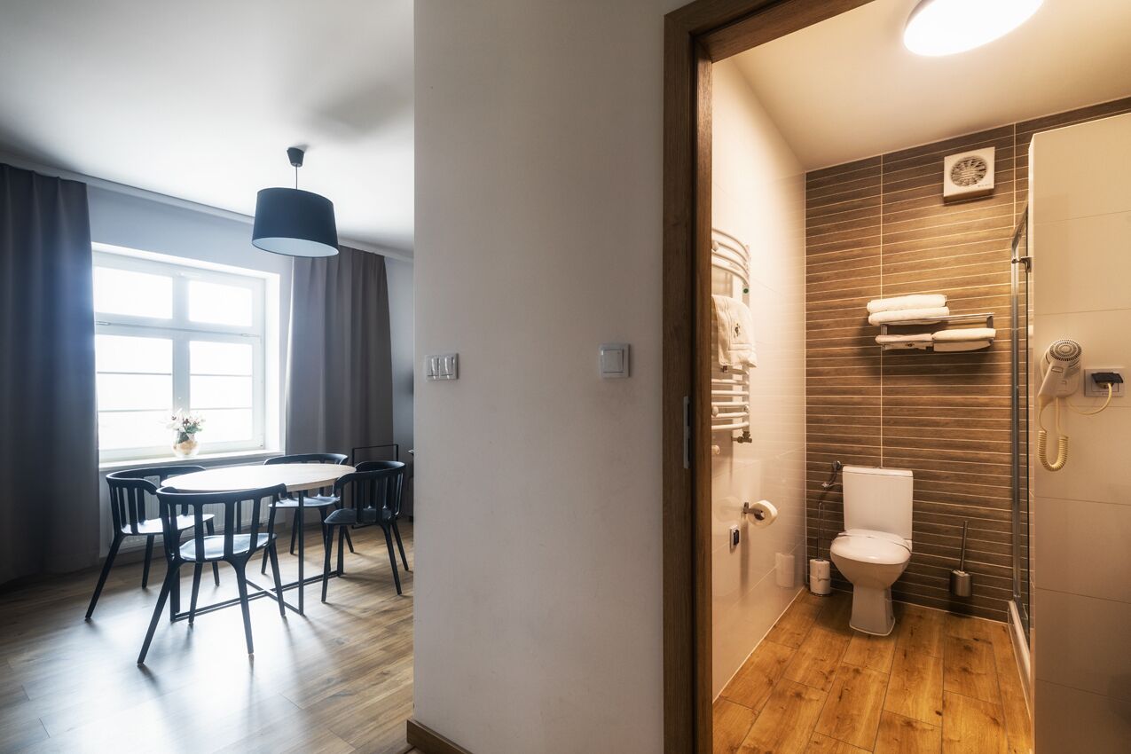 A modern hotel room with a wooden floor, a round dining table with chairs, a large window with curtains, and a bathroom with a toilet, towel rack, and shower.