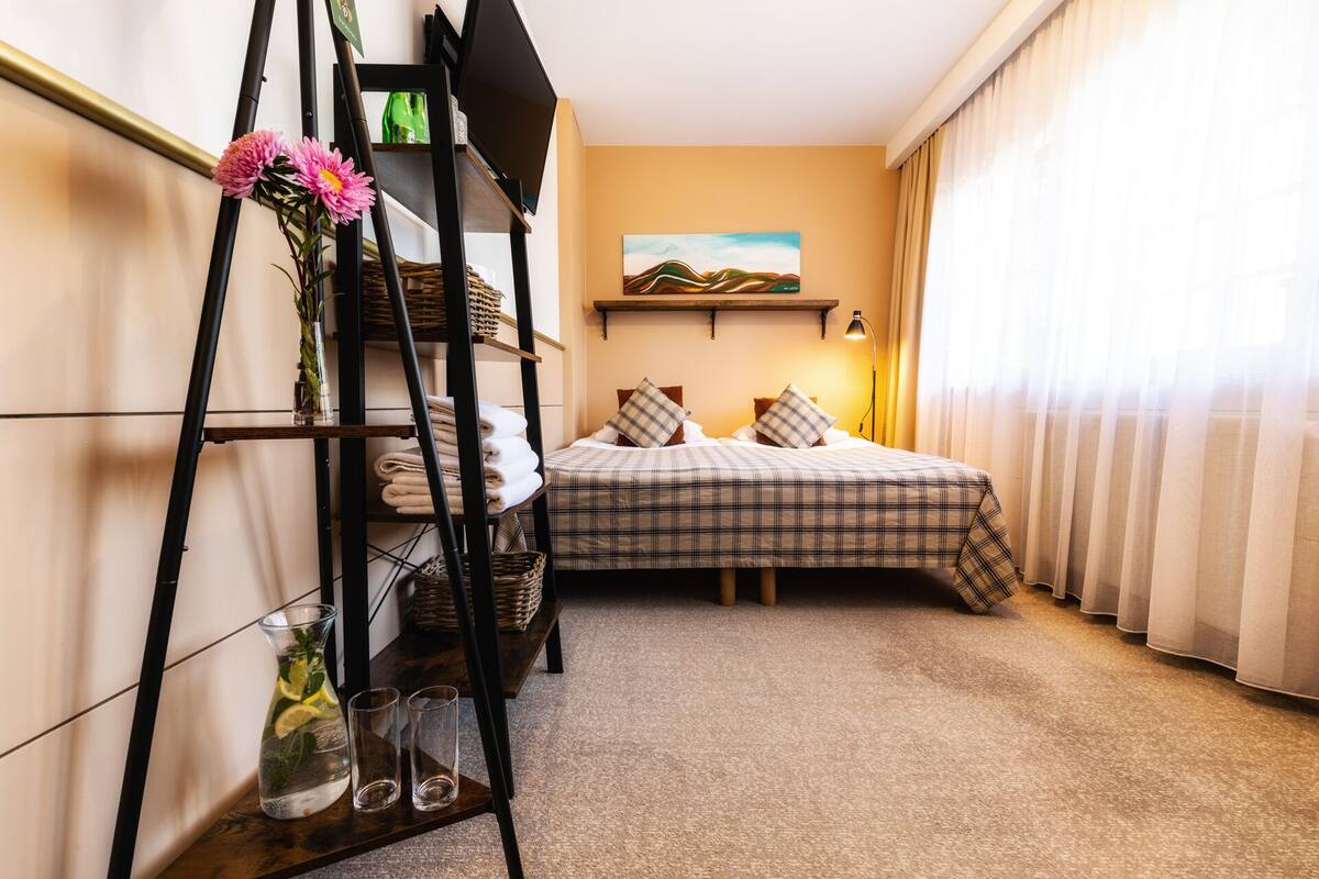 A hotel room with a bed, a shelf with towels, a vase with flowers, and a painting on the wall.
