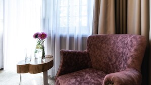 Una habitación con un sillón morado mullido, una mesita de madera con un florero y cortinas translúcidas que dejan entrar luz natural.