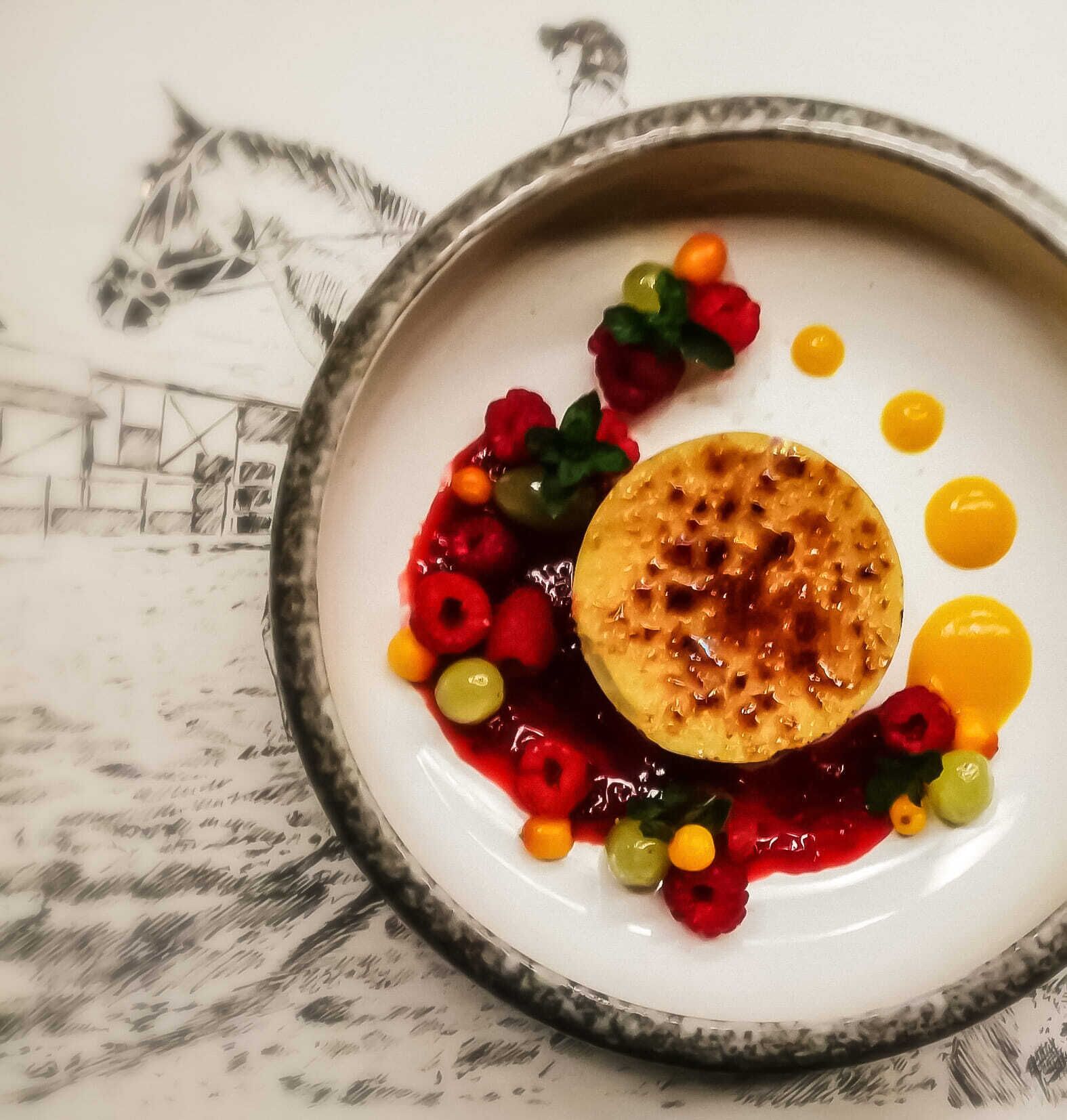 Un piatto con un dessert composto da un elemento rotondo caramellato, circondato da frutti di bosco colorati e una salsa gialla.