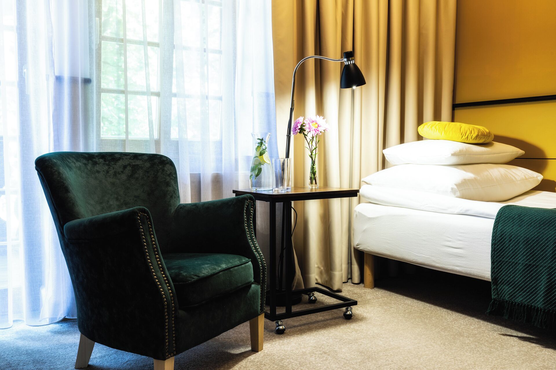 Ein Hotelzimmer mit einem grünen Sessel, einem Nachttisch mit Lampe, einer Vase mit Blumen und einem Bett mit Kissen.
