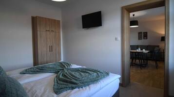 A bedroom with a bed, a wardrobe, a television, and a dining area visible through an open doorway.
