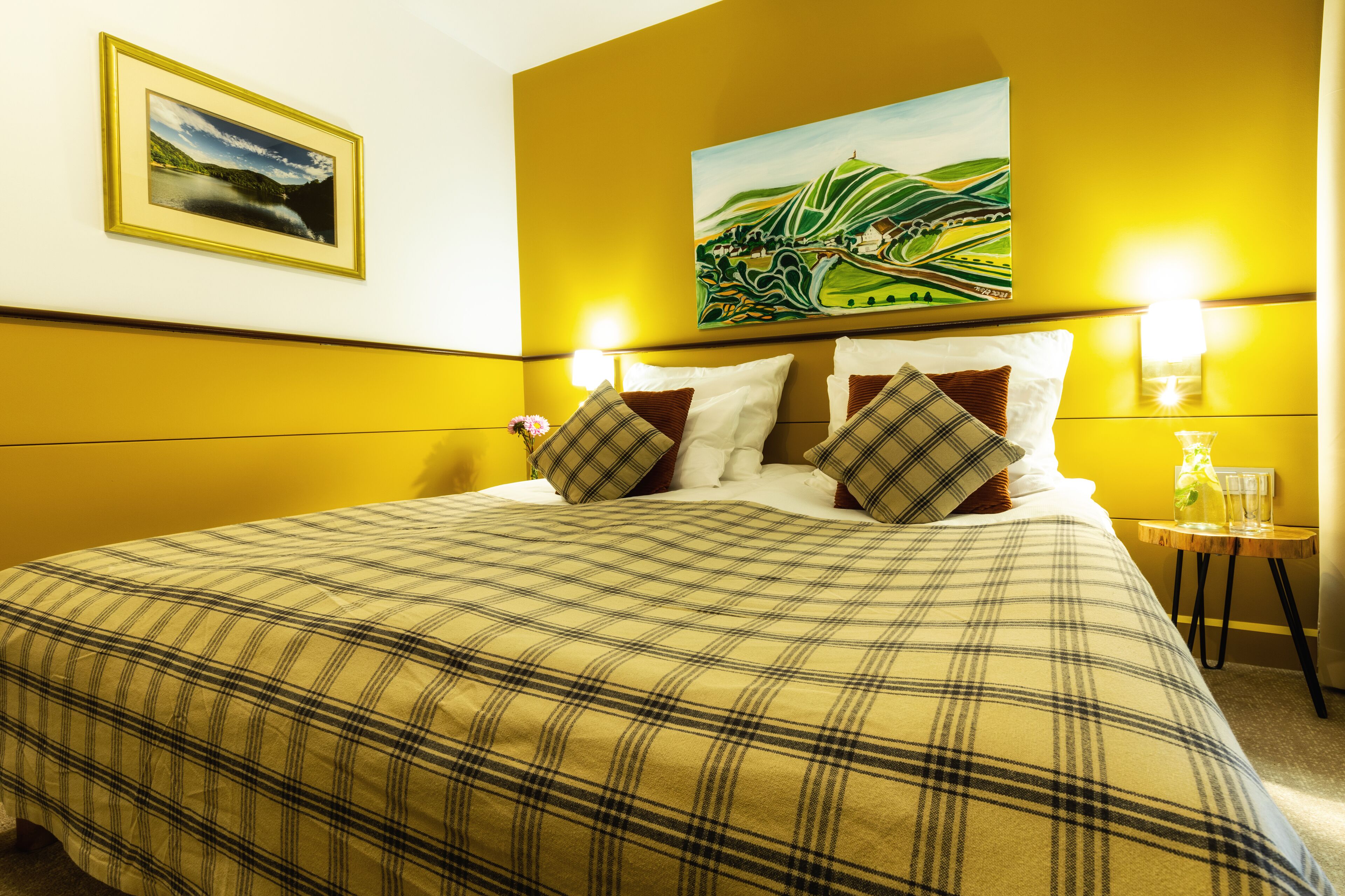 A hotel room with a large bed, a painting on the wall, and two wall-mounted lamps.