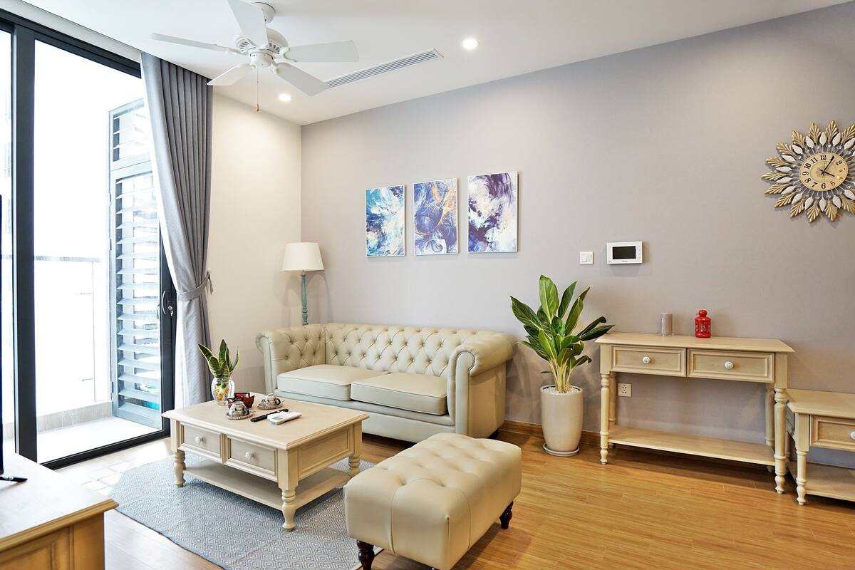 Ein modernes Wohnzimmer mit Sofa, Couchtisch, Hocker und einem Schreibtisch mit Schubladen. Es gibt einen Fernseher, einen Deckenventilator und Kunstwerke an der Wand.
