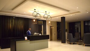 A modern reception area with a reception desk, a man standing behind it, and three hanging pendant lights. - Mythos Boutique Hotel (Kigali)