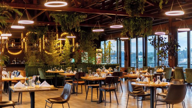 A restaurant with multiple tables and chairs, hanging plants, and large windows offering a view. - Mythos Boutique Hotel (Kigali)