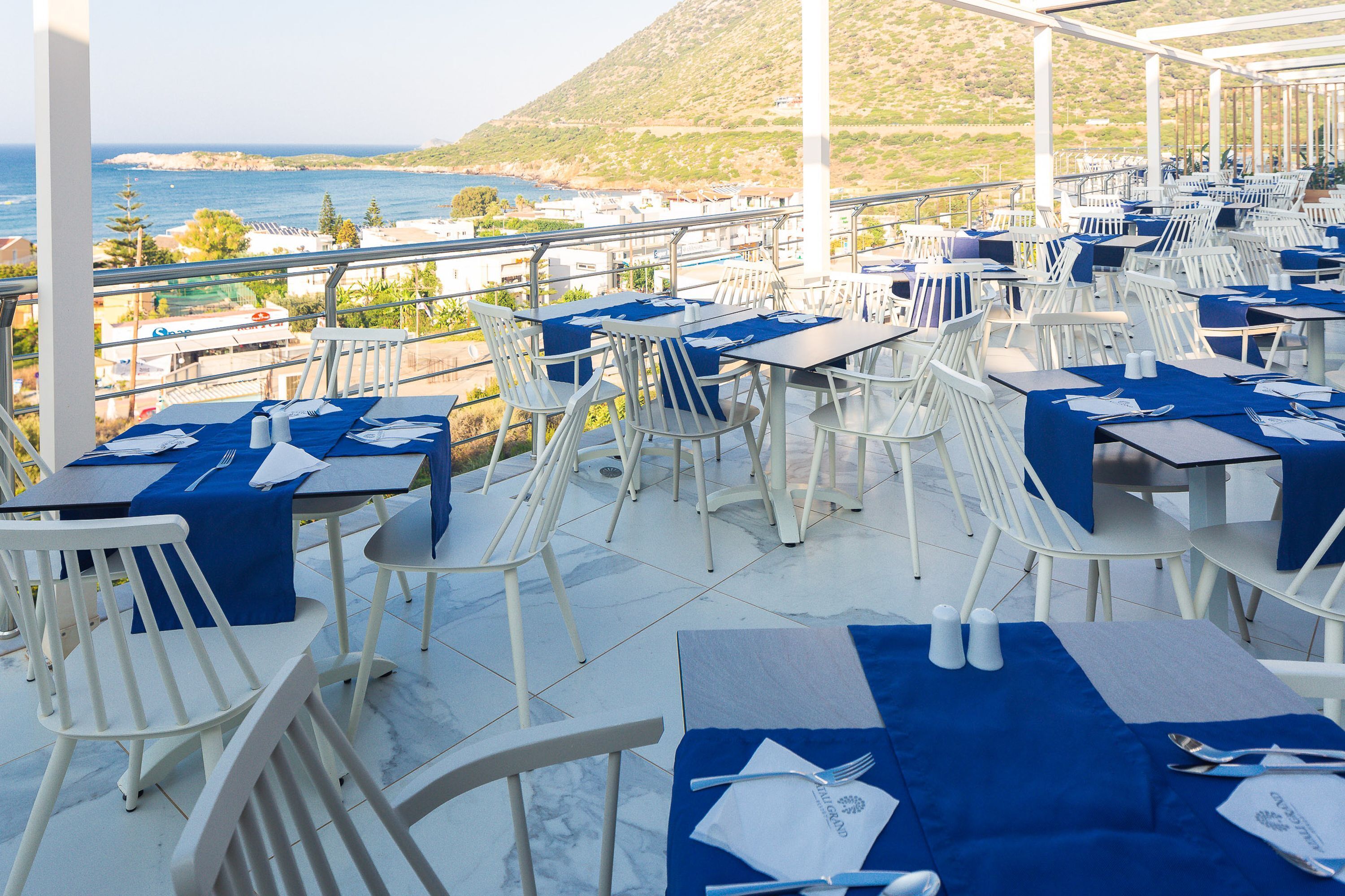 An outdoor dining area with white chairs and tables, blue tablecloths, and a view of the sea and hills.